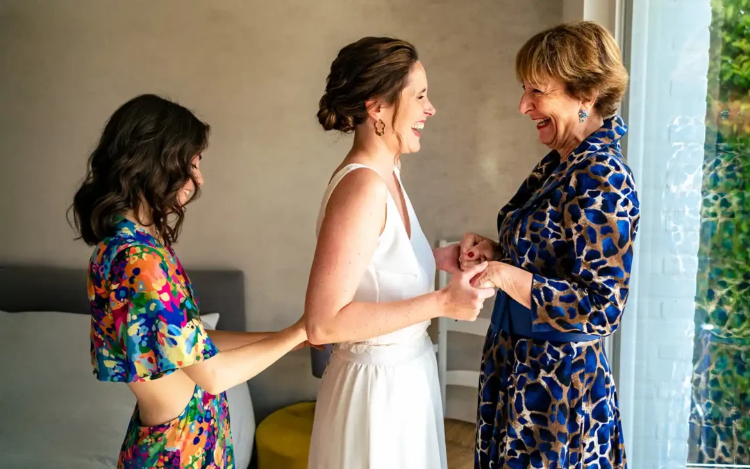 La mariée tient les mains de sa maman en souriant pendant que sa soeur ferme sa robe